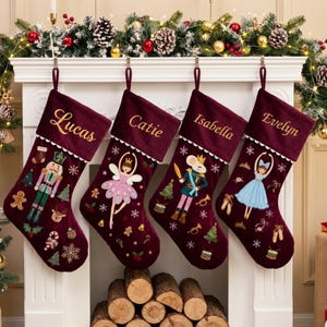 May include: Four deep red velvet Christmas stockings hanging from a white mantelpiece. Each stocking features a white cuff embroidered with the names Lucas, Catie, Isabella, and Evelyn, and displays festive motifs like nutcrackers and dancers.