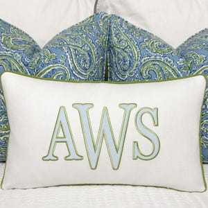 May include: A rectangular white throw pillow featuring the embroidered letters AWS in light blue with a green outline. The pillow has a green piped edge and is positioned in front of two larger pillows with a blue and green paisley pattern.