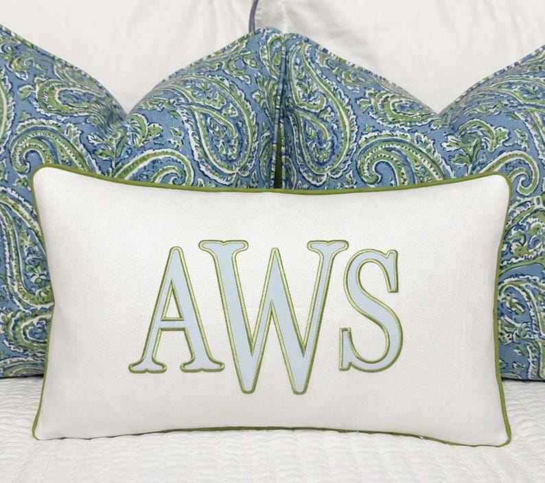 May include: A rectangular white throw pillow featuring the embroidered letters AWS in light blue with a green outline. The pillow has a green piped edge and is positioned in front of two larger pillows with a blue and green paisley pattern.