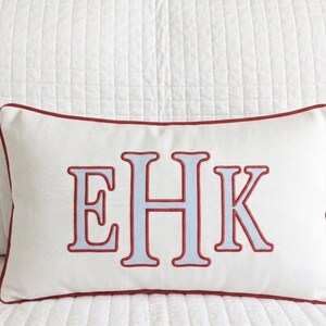 May include: A rectangular off-white throw pillow featuring a centered embroidered monogram with the letters EHK in light blue with a red outline. The pillow is finished with a red piped edge along the entire perimeter of the rectangular shape.