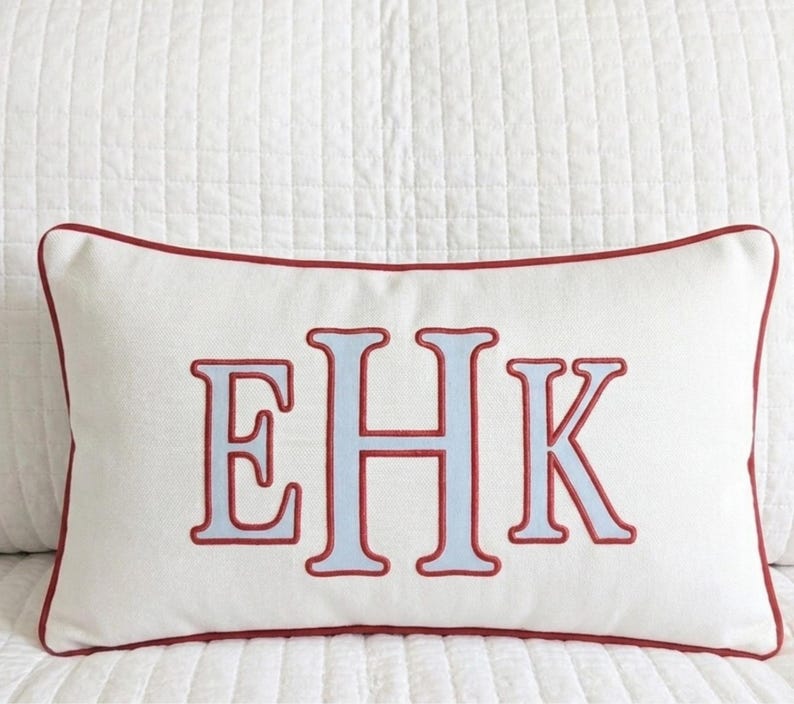 May include: A rectangular off-white throw pillow featuring a centered embroidered monogram with the letters EHK in light blue with a red outline. The pillow is finished with a red piped edge along the entire perimeter of the rectangular shape.