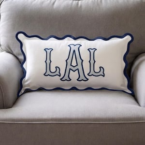 May include: A rectangular white throw pillow featuring a scalloped edge trimmed in dark navy blue. The centre of the pillow displays the embroidered letters LAL in a light blue font with a dark navy blue outline, centred on the fabric.