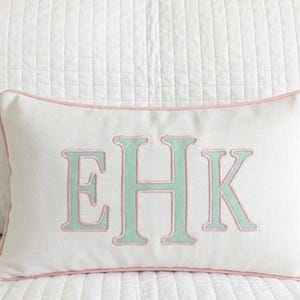 May include: A rectangular white throw pillow featuring a centered monogram with the letters EHK in light green embroidery. The letters are outlined in pink thread, and the pillow is finished with pink piping along all four edges.