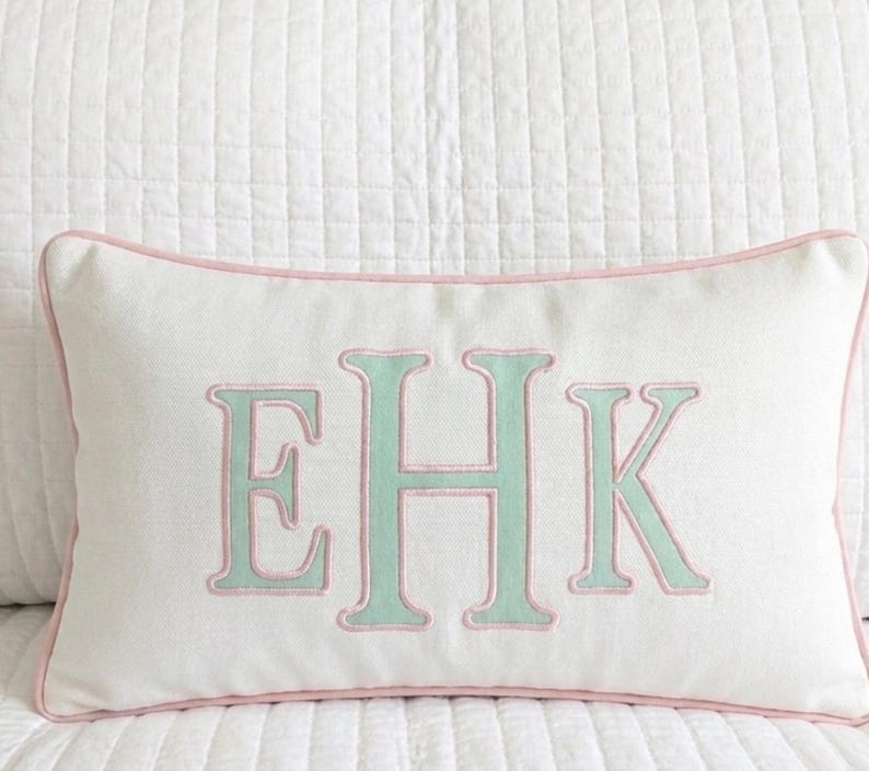 May include: A rectangular white throw pillow featuring a centered monogram with the letters EHK in light green embroidery. The letters are outlined in pink thread, and the pillow is finished with pink piping along all four edges.