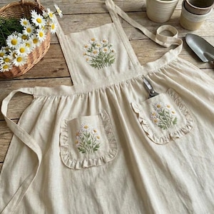 May include: A cream-coloured apron featuring embroidered daisy flowers on the pockets and bib. The apron has a ruffled edge on the pockets and a pair of scissors in one pocket. A basket of daisies and gardening tools are in the background.