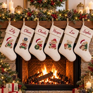 Könnte beinhalten: Sechs cremefarbene Weihnachtsstrümpfe hängen in einer Reihe, jeder mit einem Namen in roter Schreibschrift und einer einzigartigen Teddybären-Illustration bestickt. Die Bären tragen festliche Kleidung, darunter Weihnachtsmützen, Schals und Accessoires.