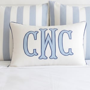 May include: A rectangular off-white throw pillow featuring a large, centered, light blue embroidered monogram with the letters CWC. The pillow is finished with a thin, dark blue piped edge, providing a clean, structured look for home decor.