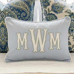May include: A rectangular light blue throw pillow featuring a white piped edge and a large, centered monogram with the letters MWM in cream-colored fabric with a light green stitched outline, centered on the front of the pillow cover.