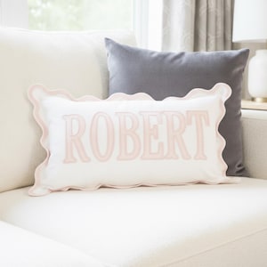 May include: A rectangular white throw pillow featuring the name ROBERT embroidered in light pink lettering. The pillow is finished with a decorative light pink scalloped edge trim that follows the wavy contour of the entire rectangular border.