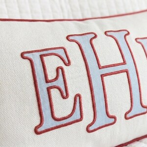 May include: A rectangular off-white throw pillow featuring large, light blue embroidered letters with a dark red stitched outline. The pillow has a matching dark red piped edge along the top seam, showcasing a textured fabric construction.
