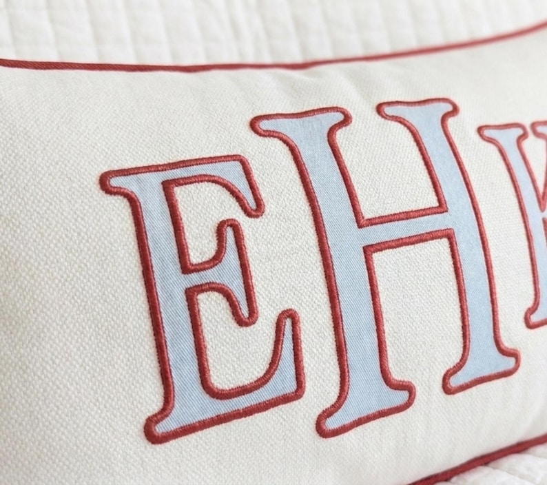 May include: A rectangular off-white throw pillow featuring large, light blue embroidered letters with a dark red stitched outline. The pillow has a matching dark red piped edge along the top seam, showcasing a textured fabric construction.