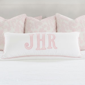 May include: A rectangular white lumbar pillow featuring the embroidered pink monogram letters JHR in the center. The pillow is finished with pink piping along the edges and is positioned in front of three square pillows with a light pink floral pattern.