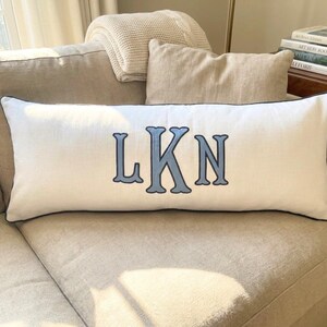 May include: A long, rectangular white lumbar pillow featuring a dark blue piped edge and the embroidered monogram letters LKN in a light blue font with a dark blue outline centered on the front of the fabric cover.
