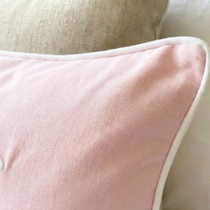 May include: A soft pink square throw pillow featuring a smooth, solid-colored fabric surface. The pillow is finished with a clean, crisp white piped edge that runs along the perimeter, providing a distinct contrast to the light pink textile.