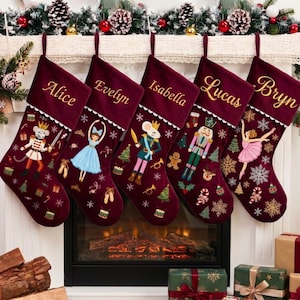 Puede incluir: Cinco calcetines navideños de terciopelo color burdeos profundo colgados en fila. Cada calcetín tiene un puño blanco bordado con un nombre: Alice, Evelyn, Isabella, Lucas o Bryn. Los calcetines están decorados con motivos de cascanueces y bailarinas.