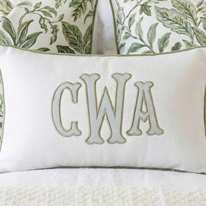 May include: A rectangular white throw pillow featuring a centered, large, light gray embroidered monogram with the letters CWA. The pillow is finished with a sage green piped edge and has a textured fabric surface, providing a clean and structured appearance.