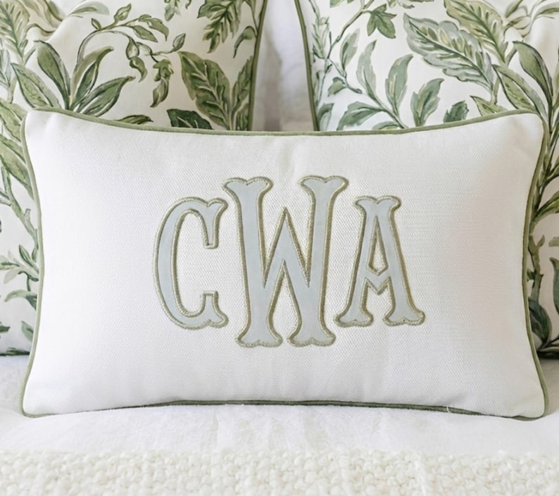 May include: A rectangular white throw pillow featuring a centered, large, light gray embroidered monogram with the letters CWA. The pillow is finished with a sage green piped edge and has a textured fabric surface, providing a clean and structured appearance.