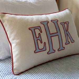 May include: A rectangular off-white throw pillow featuring the embroidered letters EHK in light blue with a dark red outline. The pillow is finished with a matching dark red piped edge along the entire perimeter of the rectangular fabric cover.