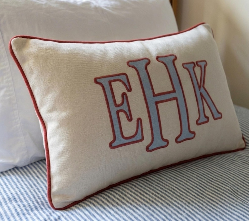 May include: A rectangular off-white throw pillow featuring the embroidered letters EHK in light blue with a dark red outline. The pillow is finished with a matching dark red piped edge along the entire perimeter of the rectangular fabric cover.