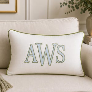 May include: A rectangular off-white throw pillow featuring the embroidered letters AWS in light blue with a sage green outline. The pillow is finished with matching sage green piping along all four edges, creating a clean and structured appearance.