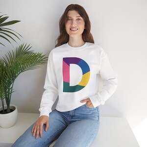 Könnte beinhalten: Weißes Sweatshirt mit einem großen, farbenfrohen Buchstaben "D" in einem geometrischen Design. Das "D" ist in verschiedenen Farben wie Lila, Rosa, Blau, Gelb und Grün unterteilt. Das Sweatshirt hat lange Ärmel und einen Rundhalsausschnitt.