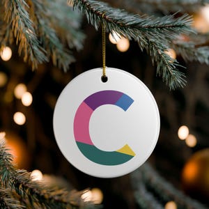 Puede incluir: Adorno navideño redondo y blanco, colgado de una rama de pino con un hilo dorado. El adorno presenta un diseño colorido de la letra "C" en morado, rosa, azul, verde y amarillo.