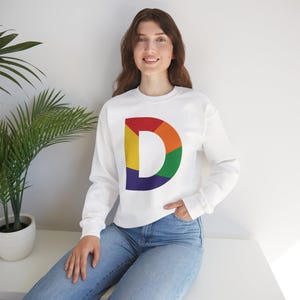 May include: White sweatshirt featuring a large, colorful letter "D" in a rainbow design. The "D" is segmented into red, orange, yellow, green, blue, and purple sections. The sweatshirt has long sleeves and a crew neck.