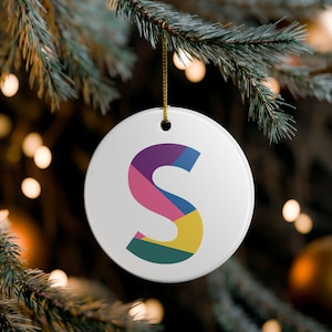 May include: White ceramic Christmas ornament with the letter "S" in a colorful geometric design. The ornament is hanging from a gold string against a blurred background of Christmas tree branches and lights.