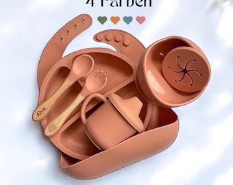 May include: A set of silicone baby feeding products in a terracotta color. The set includes a suction plate, a bowl, a cup, a spoon, and a fork. The plate has a suction base to prevent it from sliding. The set is labeled "Alva" and "4 Farben".