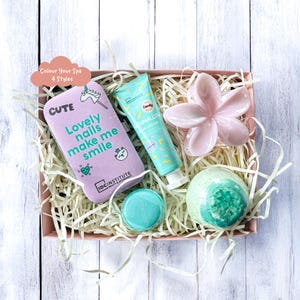Könnte beinhalten: Eine rosa Geschenkbox mit Spa-Produkten. Enthält eine lavendelfarbene Nagelfeile mit Text, eine Tube Marshmallow-Handcreme, eine rosa blütenförmige Schale, eine Badekugel und einen türkisfarbenen runden Gegenstand. Die Box ist mit Papier gefüllt.