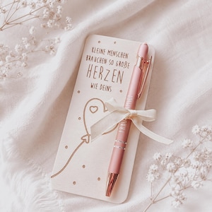Könnte beinhalten: Eine rechteckige Karte aus hellem Holz mit dem Text KLEINE MENSCHEN BRAUCHEN SO GROBE HERZEN WIE DEINS und einer herzförmigen Ballon-Illustration, kombiniert mit einem matten roségoldenen Stift, der mit einer cremefarbenen Schleife versehen ist.