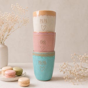 Könnte beinhalten: Ein Stapel aus drei Keramikbechern mit reaktiver Glasur. Der oberste Becher ist beige mit dem Text PAPA und einem Herz, der mittlere ist rosa mit OMA und einem Herz, und der untere ist türkis mit MAMA und einem Herz. Alle Becher sind abgerundet.