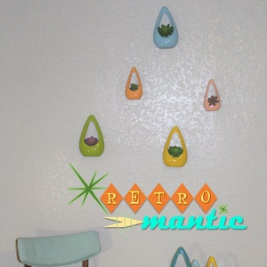 May include: Five ceramic teardrop-shaped planters in various colors hang on a white wall. The planters are arranged in a pyramid shape. The planters are filled with small plants. The word "RETRO" is written in yellow letters on a white background. The word "mantic" is written in blue letters on a white background.