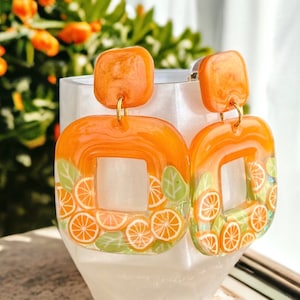 Puede incluir: Un par de pendientes naranjas y verdes con un diseño cuadrado y un gancho dorado. Los pendientes están hechos de resina y presentan un diseño de cítricos.