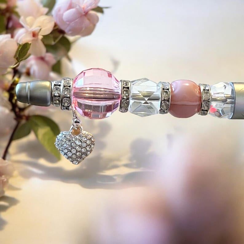 May include: A pink and silver pen with a heart-shaped charm. The pen is decorated with rhinestones and beads.