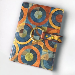 May include: A brown leather notebook with a geometric pattern of circles and squares in shades of blue, orange, and yellow. The notebook has a gold metal ring and a brown leather strap with gold metal studs.