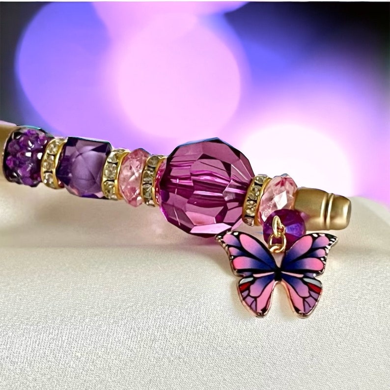 May include: A purple and pink beaded pen with a gold-toned tip and a pink butterfly charm. The pen is decorated with clear rhinestones and a large purple crystal bead.