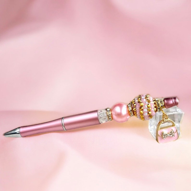 K&ouml;nnte beinhalten: Ein rosa Stift mit einer Perlen- und Goldperlenverzierung und einem rosa Handtaschenanh&auml;nger. Der Stift liegt auf einem rosa Hintergrund.