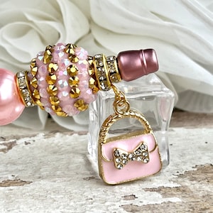 K&ouml;nnte beinhalten: Ein rosa und goldenes Charm-Armband mit einem rosa Handtaschen-Anh&auml;nger. Der Handtaschen-Anh&auml;nger hat eine goldene Kette und eine goldene Schleife mit Strasssteinen.