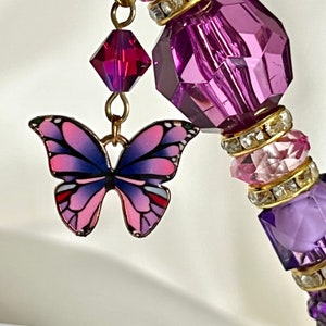 May include: A purple and pink beaded charm with a gold chain and a butterfly charm. The butterfly charm is pink with purple wings and a gold accent.