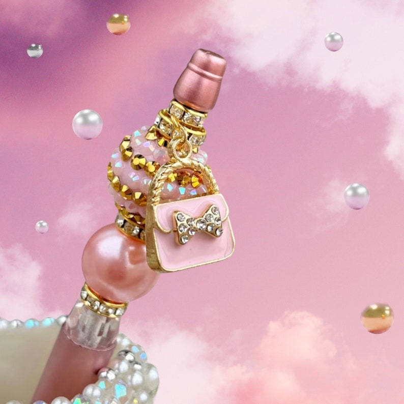 K&ouml;nnte beinhalten: Ein rosa und goldener dekorativer Flaschenverschluss mit einem kleinen rosa Handtaschen-Anh&auml;nger. Der Handtaschen-Anh&auml;nger hat eine goldene Kette und eine goldene Schleife mit Strasssteinen. Der Flaschenverschluss ist mit Strasssteinen und Perlen verziert.