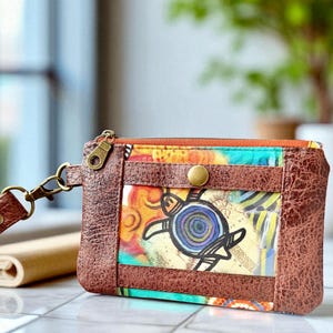 May include: A small, brown leather wallet with a colorful turtle design. The wallet features a zipper closure, a wrist strap, and a button clasp. The design includes a turtle illustration with blue, yellow, and green accents, set against a map-like background.