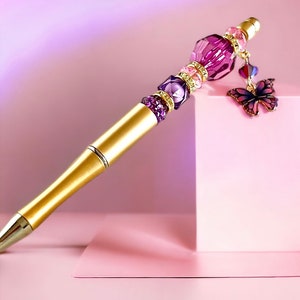 May include: A gold pen with a purple and pink crystal embellishment and a butterfly charm. The pen is on a pink surface.