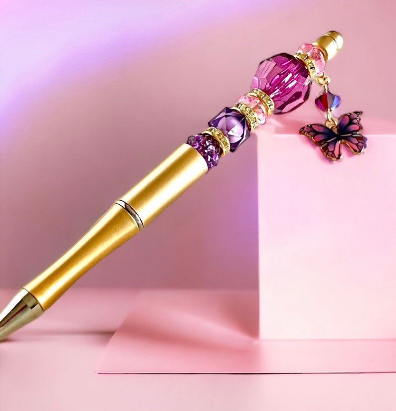 May include: A gold pen with a purple and pink crystal embellishment and a butterfly charm. The pen is on a pink surface.