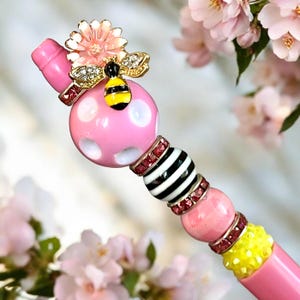 May include: A decorative pen with a pink and white polka dot bead, adorned with a gold bee and flower embellishment. The pen features black and white striped beads, pink and yellow accents, and red rhinestone details. The pen is set against a soft floral background.