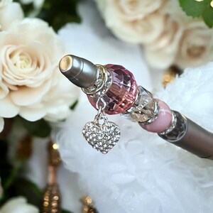 May include: A silver pen with a pink and clear crystal embellishment and a silver heart charm with clear rhinestones.