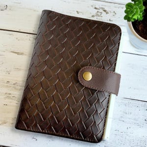 May include: A brown leather planner with a woven pattern and a gold button closure. The planner has a white interior.