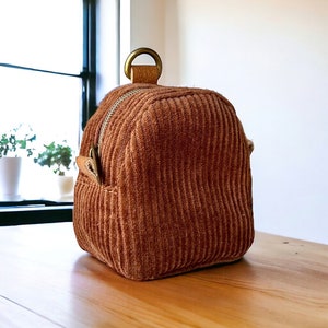 May include: A small, brown corduroy bag with a zipper closure and a brass loop handle. The bag has a textured, ribbed surface.