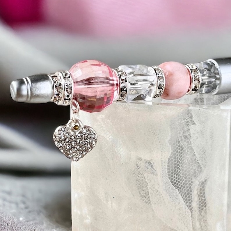 May include: A silver pen with a pink and clear crystal bead design. The pen has a heart-shaped charm with rhinestones.