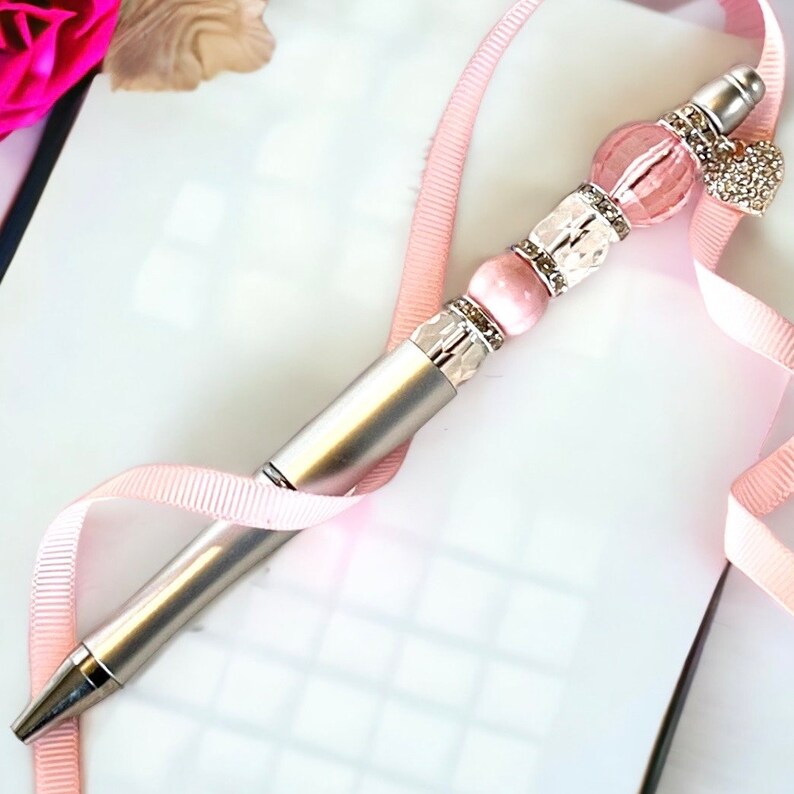 May include: A silver pen with a pink and white beaded design. The beads are accented with rhinestones and a small heart charm.